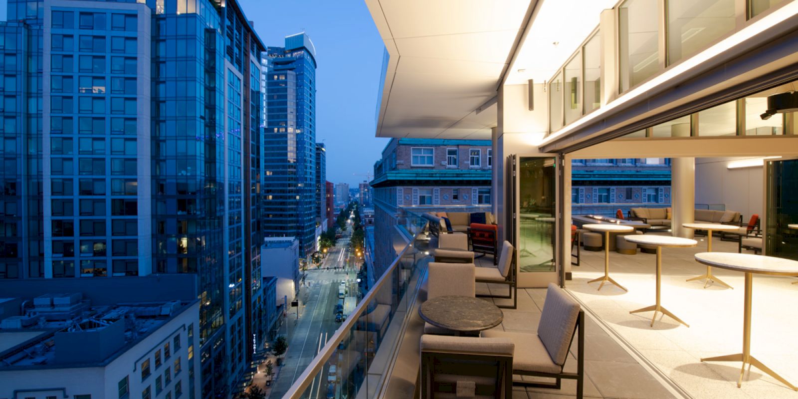 A modern balcony overlooking a city street at dusk, with outdoor seating and tables, glass railings, and high-rise buildings in the background.