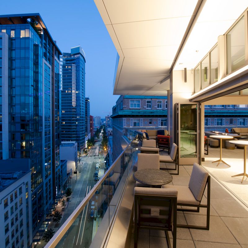 A modern cityscape view from a balcony at dusk, featuring outdoor seating and tables with nearby tall buildings lightly illuminated.