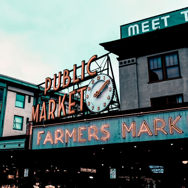 The image shows an outdoor farmers market with neon signs for "Public Market" and "Farmers Market," along with surrounding buildings and a clock.