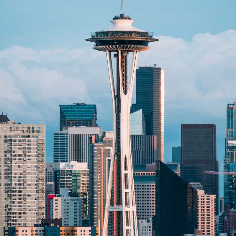 The image shows a cityscape with the Space Needle in the foreground, surrounded by modern skyscrapers against a partly cloudy sky.