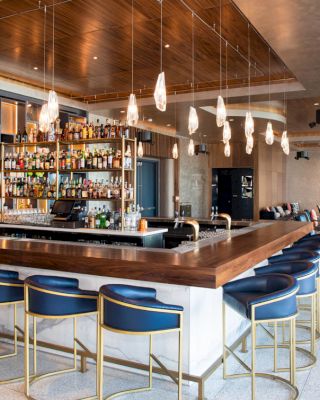 The image shows a modern bar with blue barstools, a stocked back bar, pendant lights, and seating areas in the background.