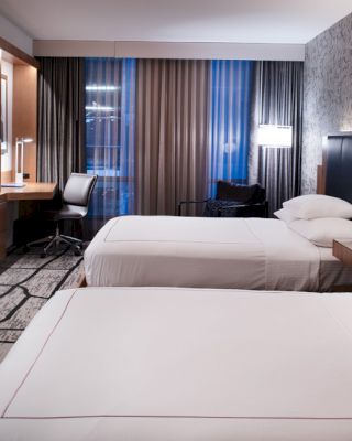 The image shows a modern hotel room with two neatly made beds, a wall-mounted TV, a desk with a chair, and stylish decor.