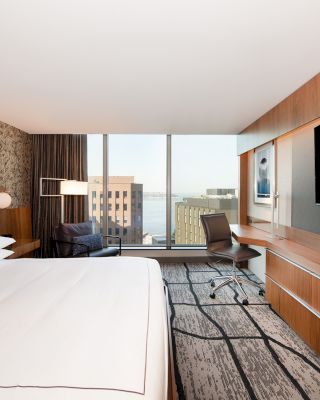 A modern hotel room with a large bed, a desk, a chair, a TV, and a view of buildings and water through floor-to-ceiling windows.