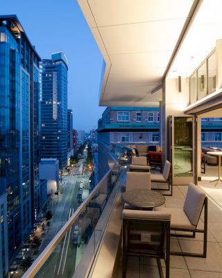 A modern rooftop dining area with tables and chairs overlooks a cityscape of tall buildings in the evening, with lights on in the buildings.