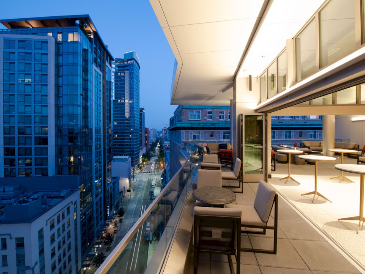 An urban balcony with tables and chairs overlooks a cityscape of tall buildings during dusk, with lights illuminating the street below.