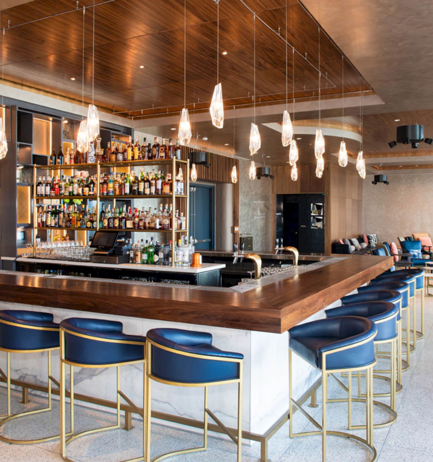 A modern bar with a wooden counter, blue cushioned chairs, pendant lights, and shelves filled with various bottles of alcohol.