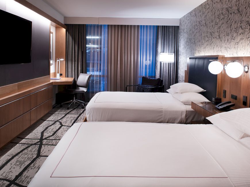 This image shows a modern hotel room with two beds, a TV, a desk with a chair, nightstands, lamps, and large windows with curtains.