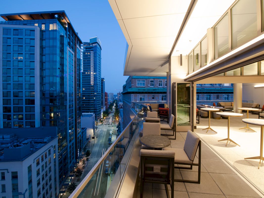 A modern cityscape at dusk with high-rise buildings; a spacious balcony includes seating and tables, providing a view of the bustling street below.