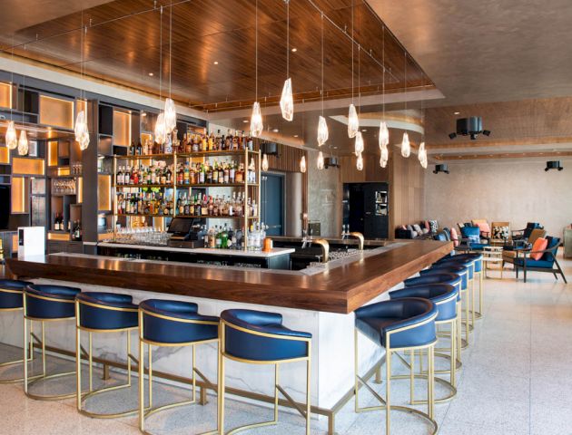 The image shows a modern bar with a wooden counter, blue bar stools, a fully stocked bar, pendant lighting, and a lounge area in the background.