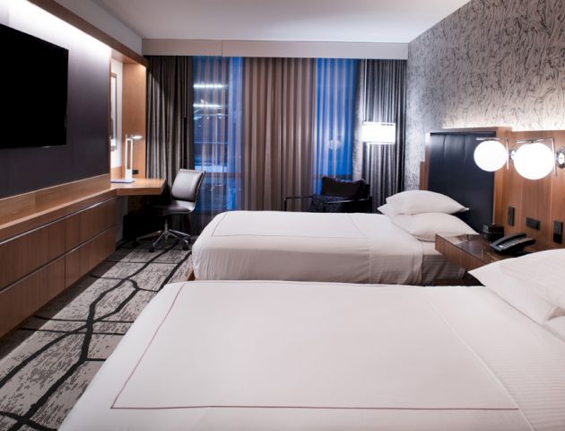 A modern hotel room with two beds, a desk, a TV, and a stylish decor featuring patterned wallpaper and a large window with curtains.