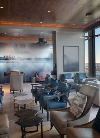The image showcases a modern lounge with comfortable seating, large windows offering a waterfront view, and a cityscape mural on the back wall.