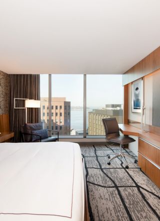 A modern hotel room with a large bed, a desk and chair, wall-mounted TV, and large windows offering a city and water view, featuring minimalist decor.