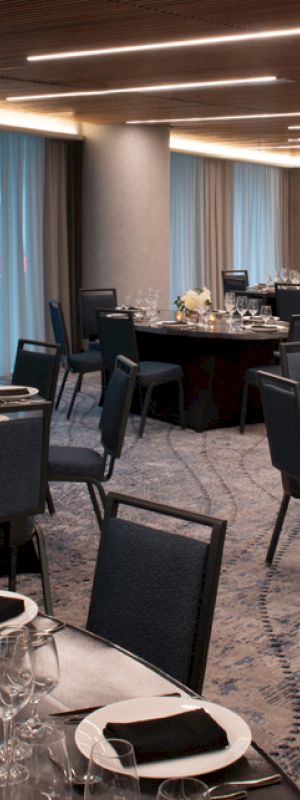 The image shows an elegant dining room set up with round tables, black chairs, neatly arranged table settings, and soft lighting.