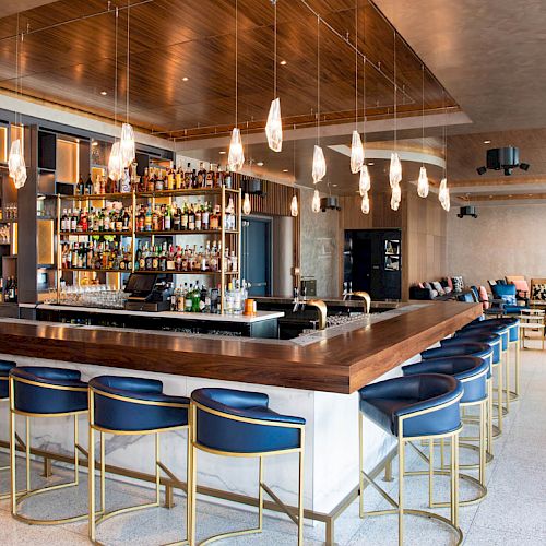 A modern bar with a wooden counter, blue bar stools, hanging lights, and shelves stocked with various bottles. The area includes a cozy seating area.