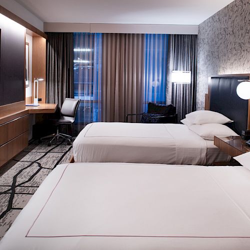 A modern hotel room with two neatly made beds, a wall-mounted TV, a desk, and contemporary decor lit by soft, ambient lighting.