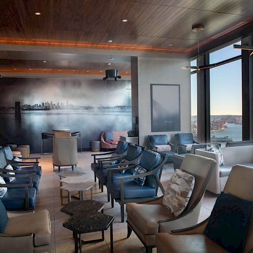 A neatly arranged lounge area with comfortable chairs, tables, a wall mural, and large windows offering a scenic waterfront view with a Ferris wheel.