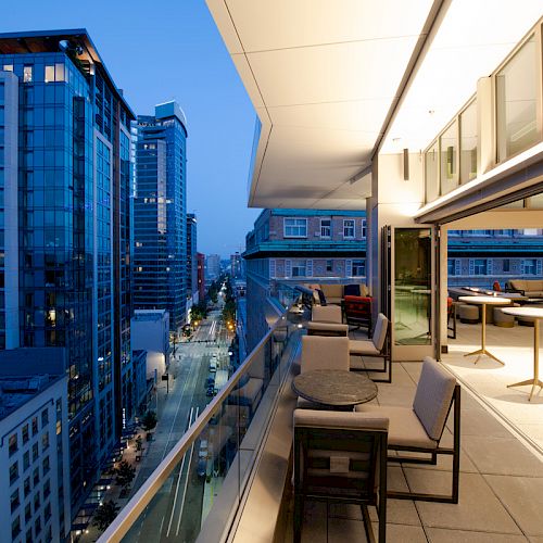 A modern cityscape at dusk with a high-rise balcony featuring outdoor seating and tables, overlooking a street lined with tall buildings.