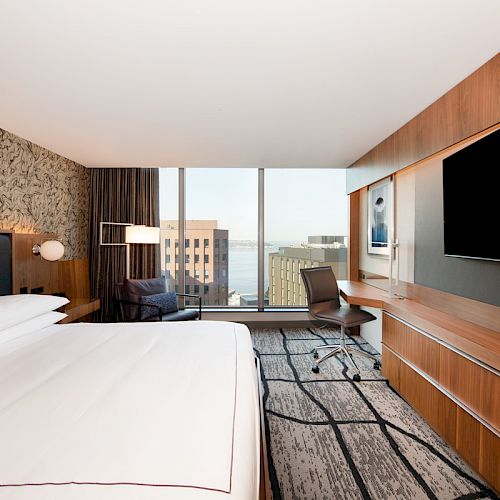 A modern hotel room with a large bed, desk, chair, and a flat-screen TV; large windows offer a cityscape view.