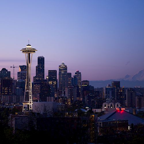 The image depicts the Seattle skyline at dusk, featuring the iconic Space Needle and Mount Rainier in the background, with city lights starting to glow.