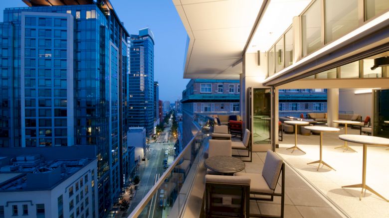 A modern cityscape at dusk with high-rise buildings and a sophisticated, well-lit balcony featuring tables and seating overlooking the urban view.