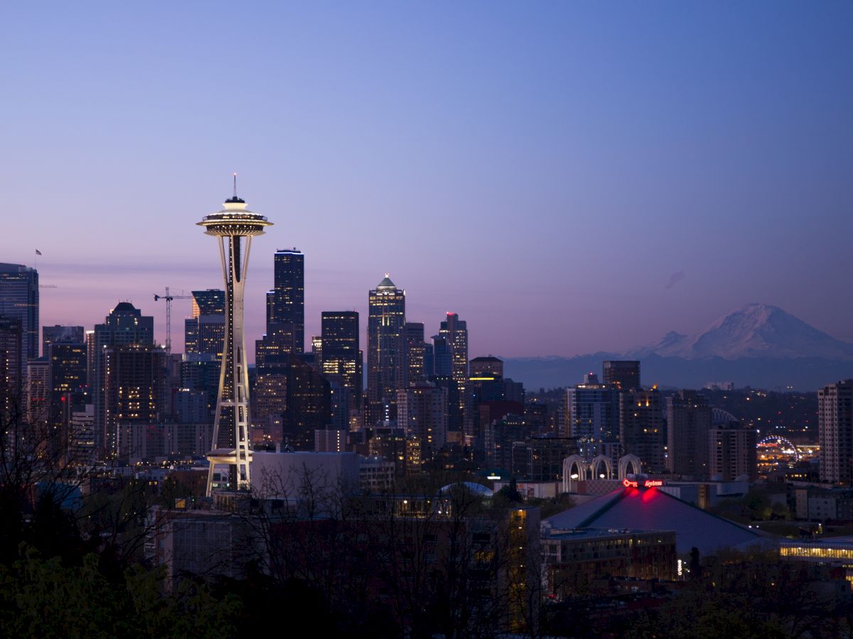 A view of Seattle's skyline at dusk, featuring the Space Needle prominently with Mount Rainier visible in the background, and city lights beginning to sparkle.