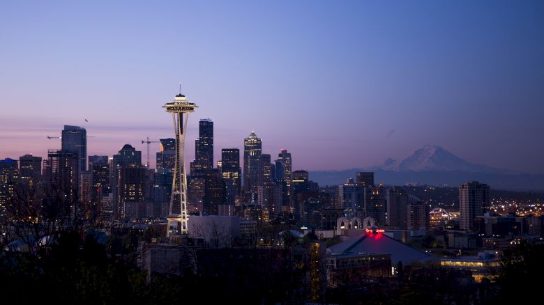 A view of Seattle's skyline at dusk, featuring the Space Needle prominently with Mount Rainier visible in the background, and city lights beginning to sparkle.