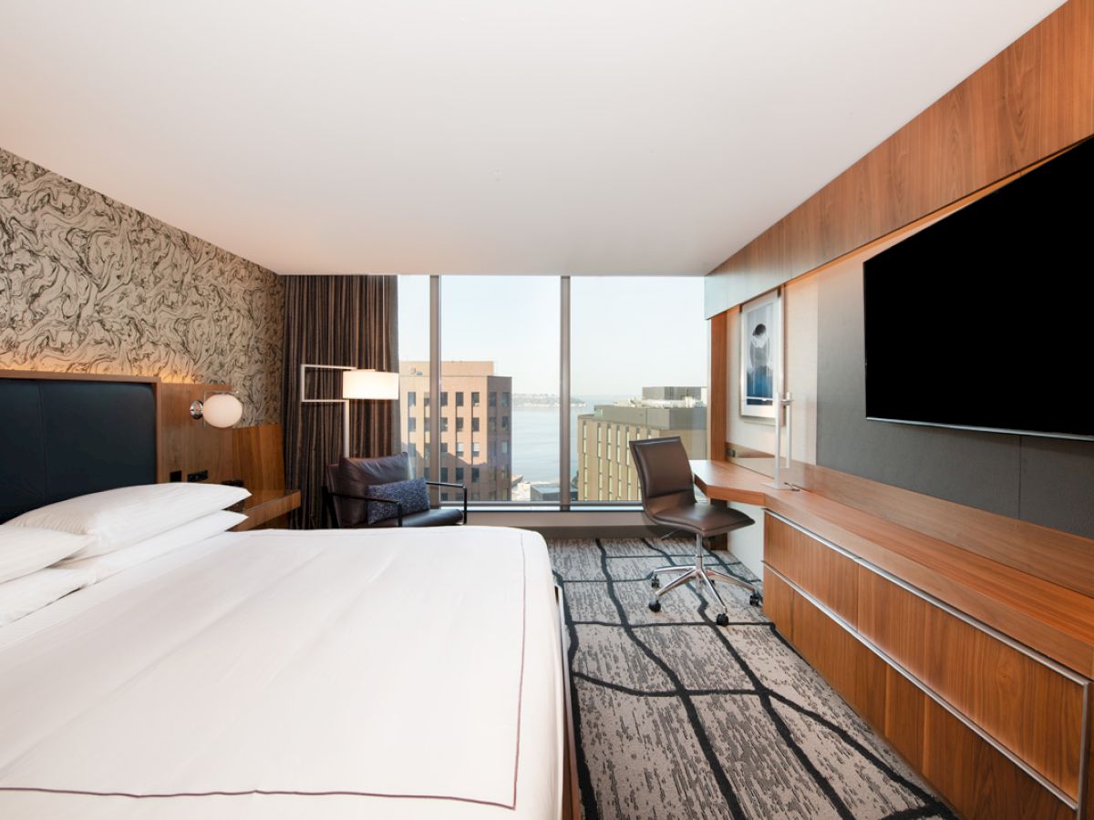 A modern hotel room with a large bed, television, desk, chair, and expansive windows offering a view of buildings and water outside.
