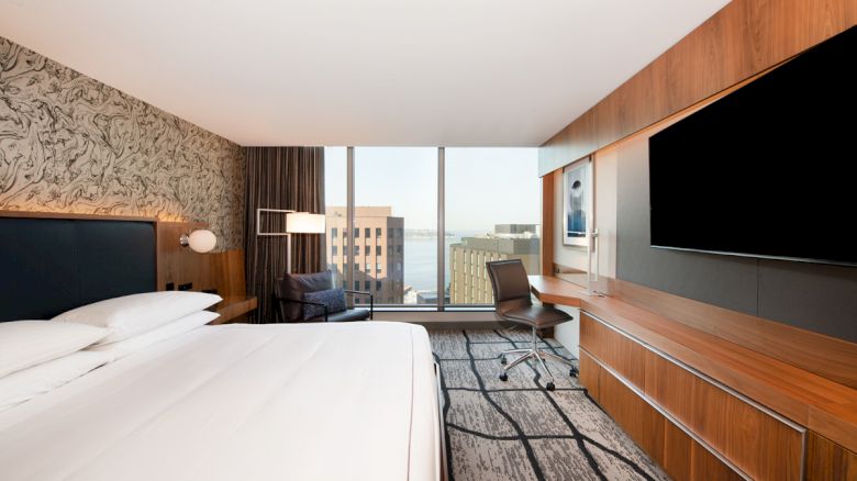 A modern hotel room with a large bed, television, desk, chair, and expansive windows offering a view of buildings and water outside.
