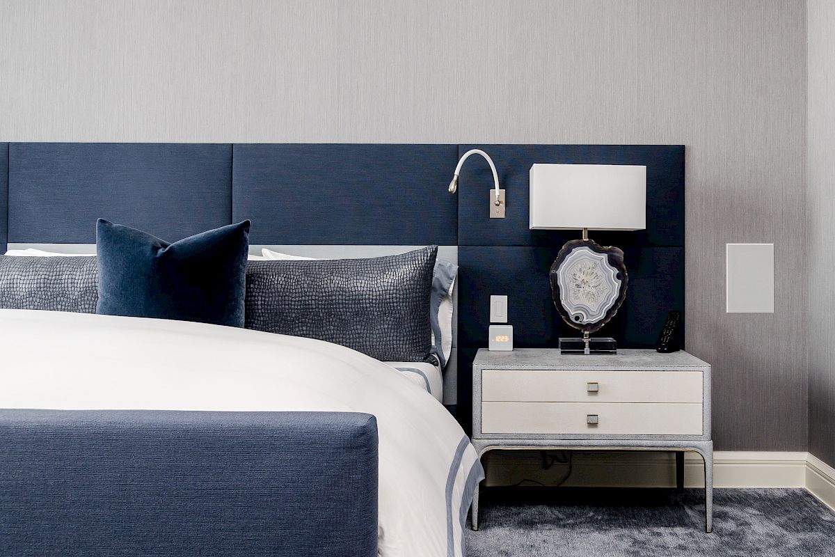A stylish bedroom with a blue and white color scheme, featuring a blue upholstered headboard, nightstand with lamp, and neatly made bed.