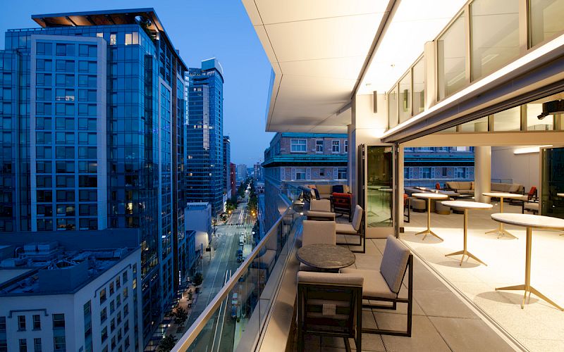 A modern outdoor terrace with seating and tables overlooks a cityscape of tall buildings at dusk, under a clear evening sky.