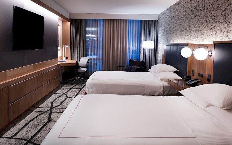A modern hotel room with two beds, a wall-mounted TV, a desk with a chair, and contemporary lighting and decor.