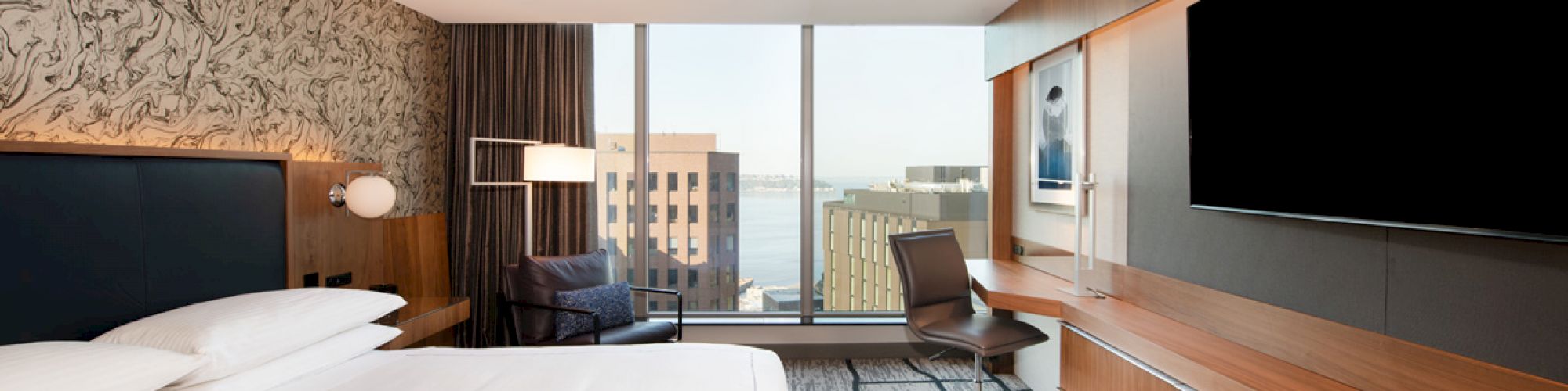 A modern hotel room with a large bed, wall-mounted TV, desk, chair, and window showing a cityscape and water view in the background.
