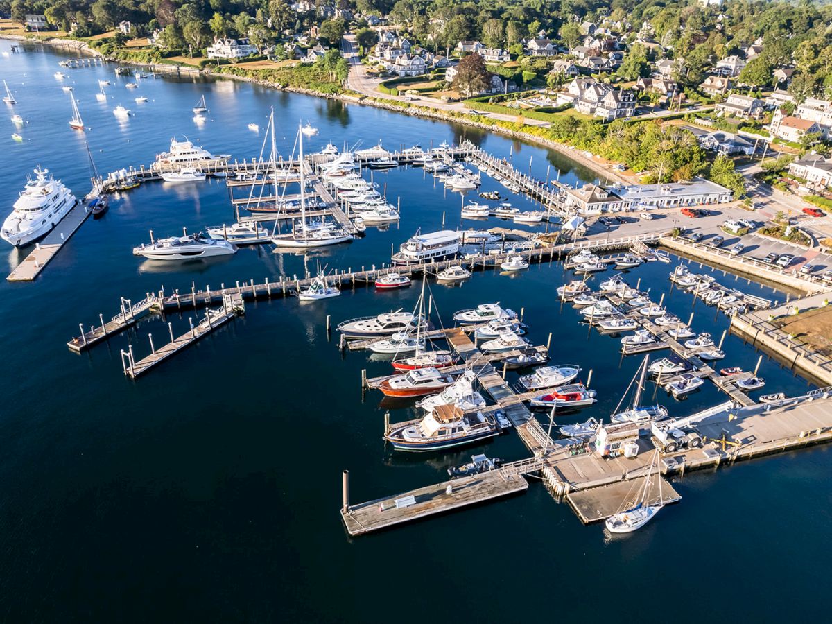 An aerial view of a marina with numerous boats docked, surrounded by calm water and nearby residential buildings and greenery.