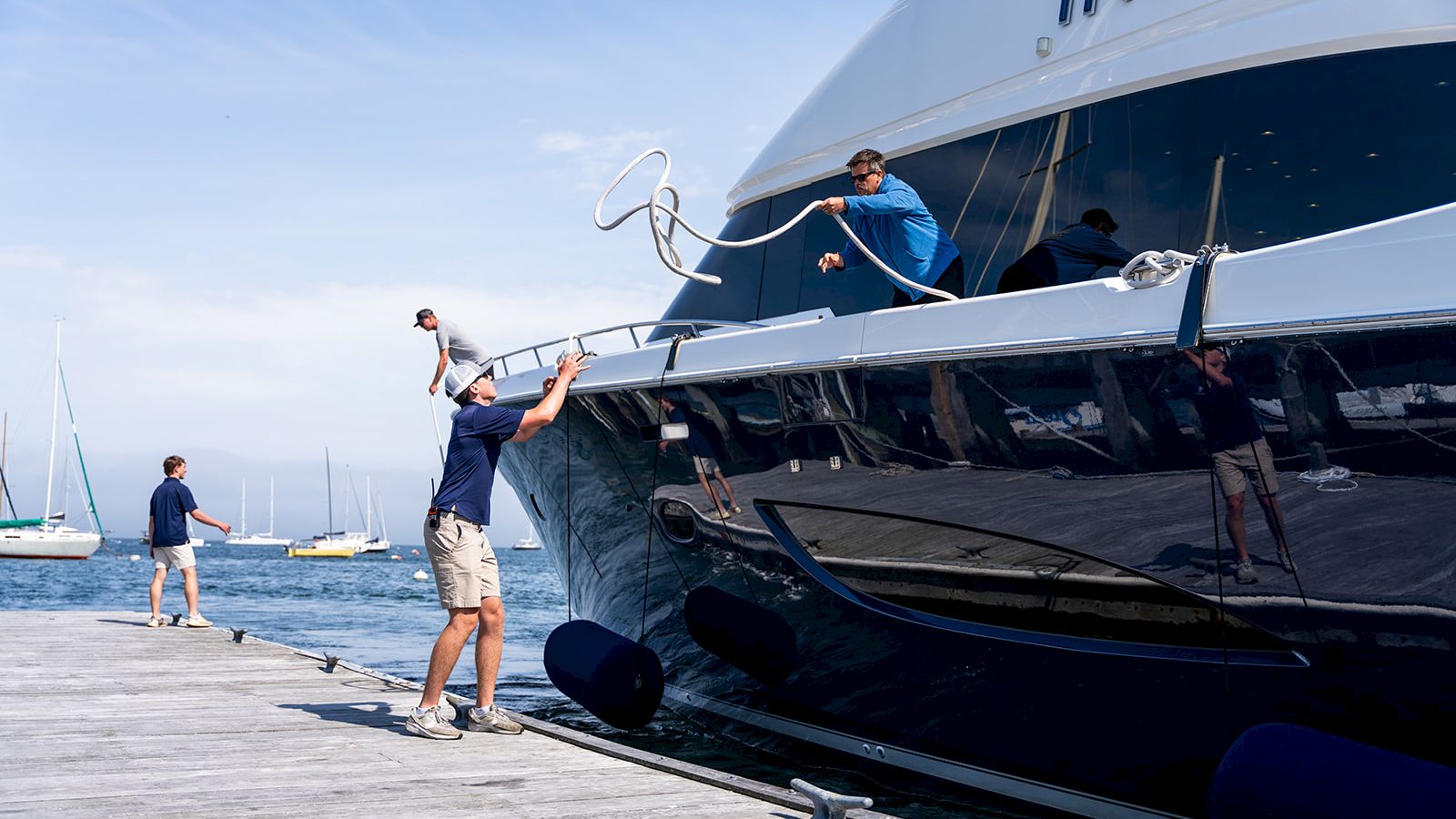 Two men lift a heavy rope onto a large white yacht at a marina, while a third man stands on the dock watching.
